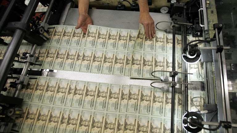 A worker's hands hover over recently printed twenty-dollar bills, inspecting them.