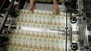 A worker's hands hover over recently printed twenty-dollar bills, inspecting them.