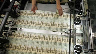 A worker's hands hover over recently printed twenty-dollar bills, inspecting them.