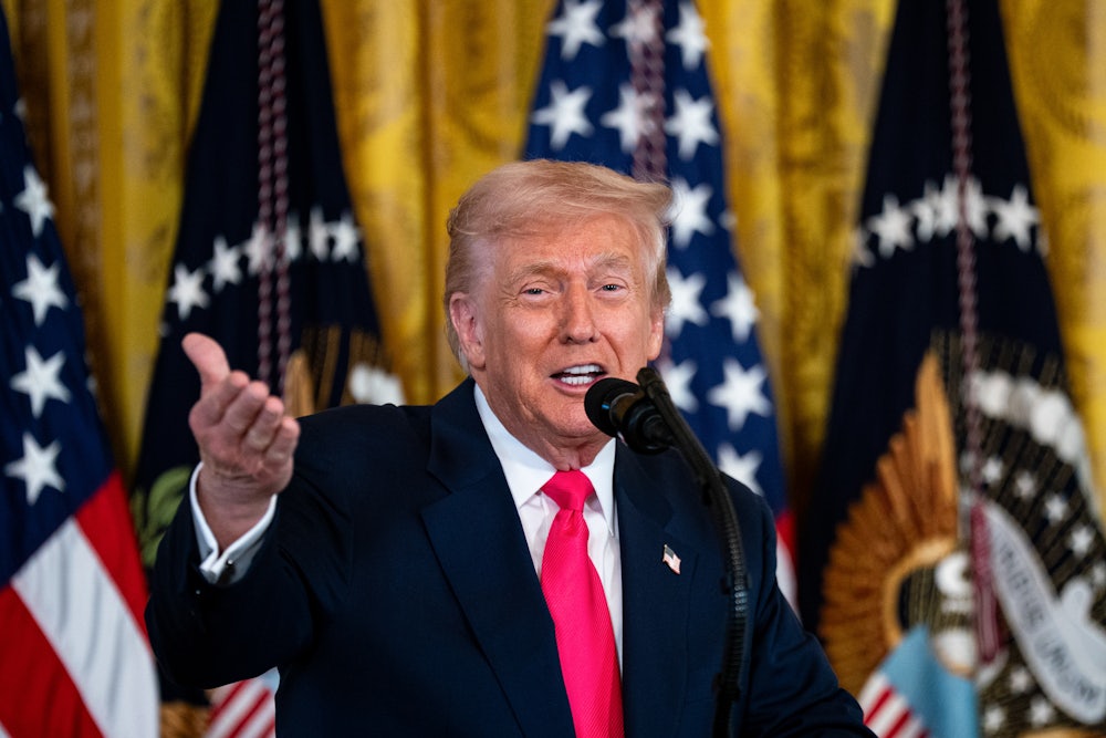 Trump gestures with one hand while speaking into a microphone, standing in front of flags.