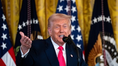 Trump gestures with one hand while speaking into a microphone, standing in front of flags.