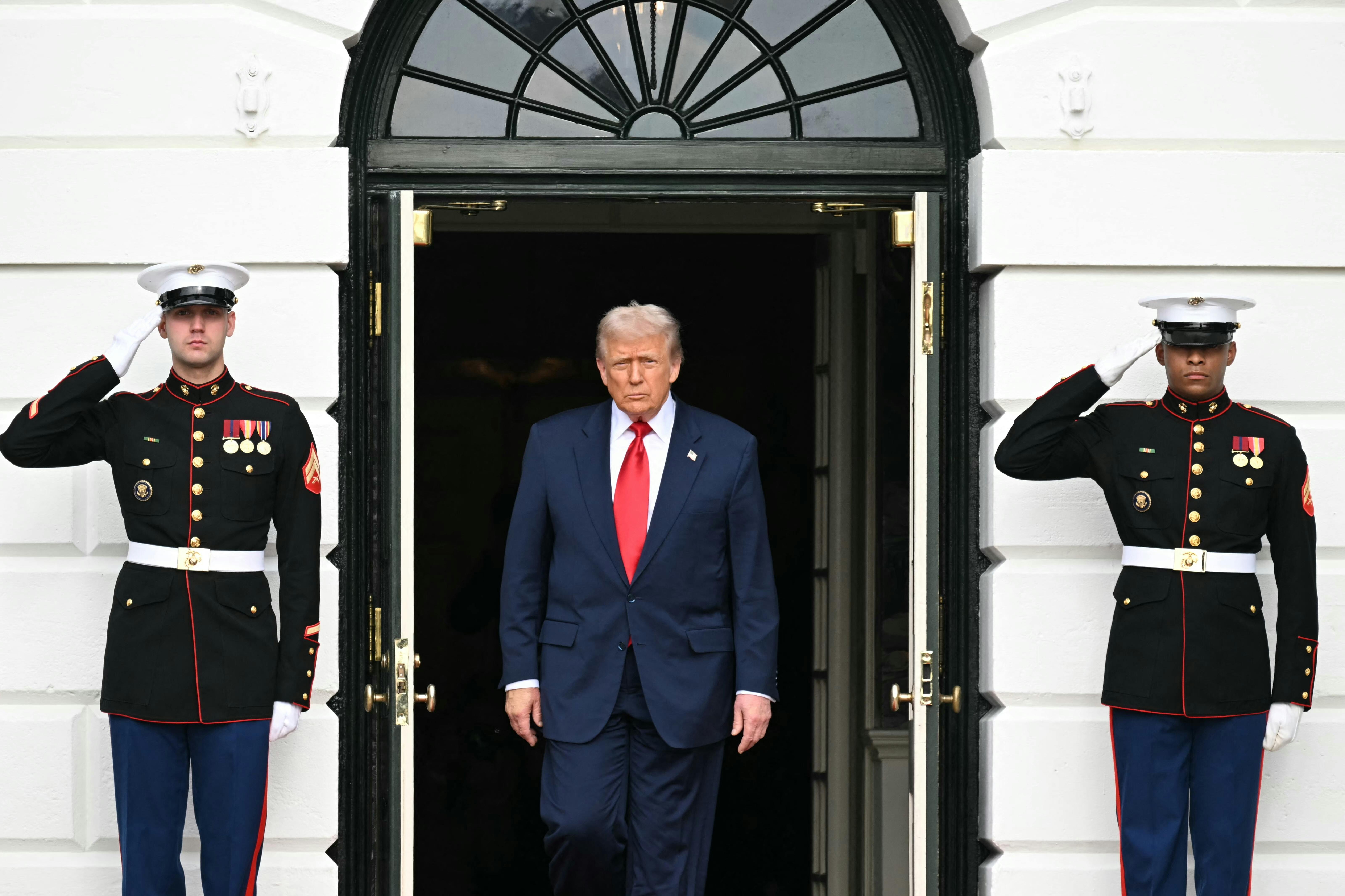 Donald Trump walks out of a door of the White House. The door is flanked on each side by a saluting soldier