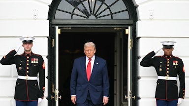 Donald Trump walks out of a door of the White House. The door is flanked on each side by a saluting soldier