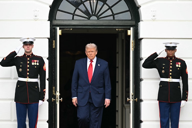 Donald Trump walks out of a door of the White House. The door is flanked on each side by a saluting soldier