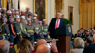 Trump stands at a podium and twists to look behind him at rows of men in hard hats.