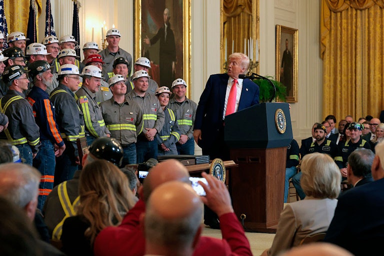 Trump stands at a podium and twists to look behind him at rows of men in hard hats.