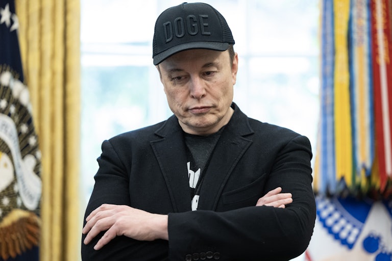 Elon Musk wears a black DOGE cap and crosses his arms while looking down. He is in the Oval Office of the White House.