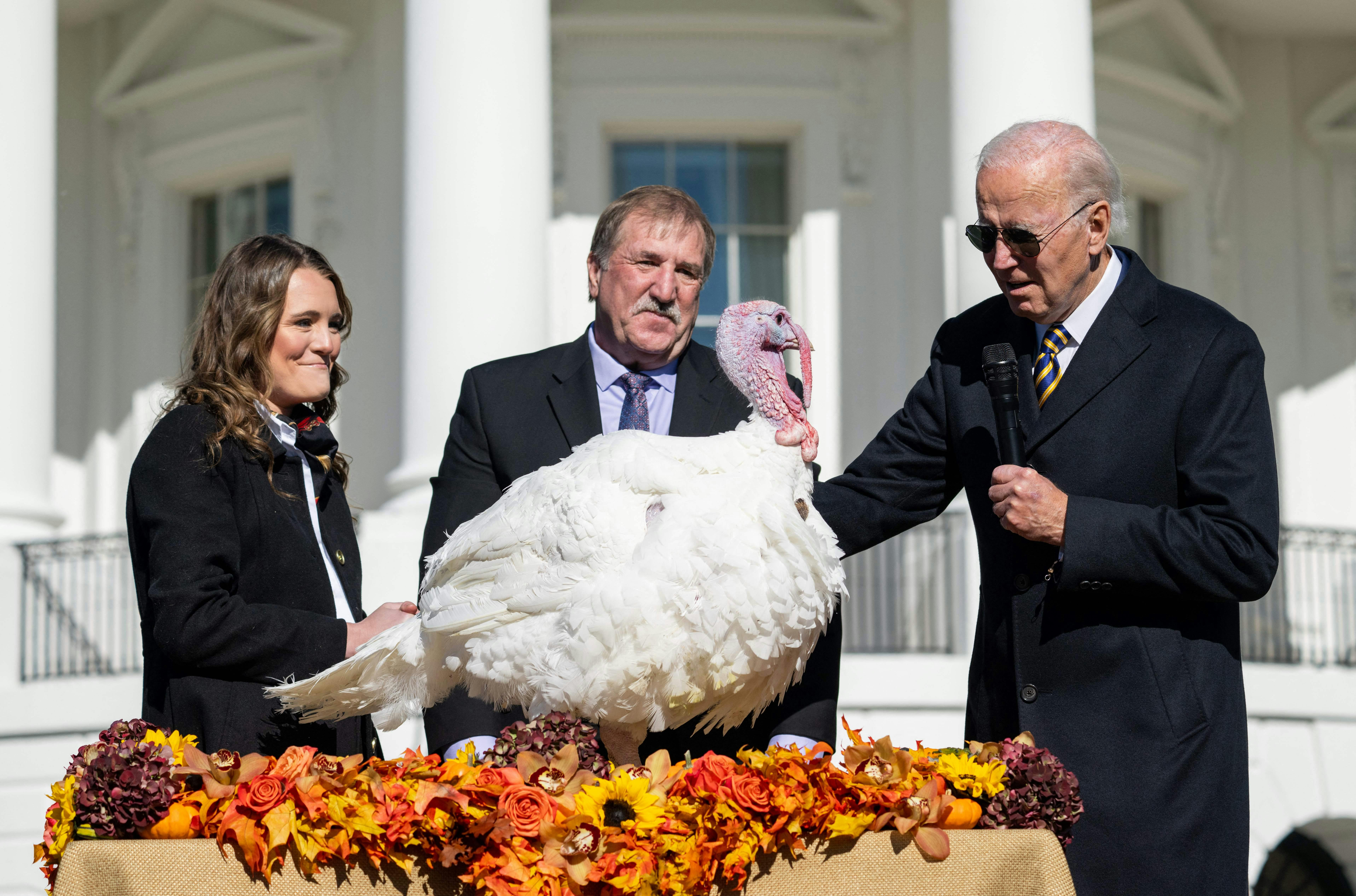Joe Biden and two others stand behind a turkey positioned on a table festooned with autumn leaves and flowers.