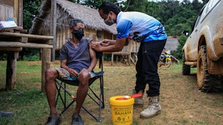 A health worker injects a Covid vaccine into a seated man's arm, outdoors, in front of a hut.
