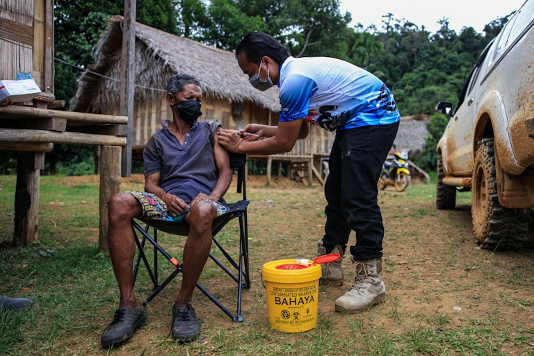 A health worker injects a Covid vaccine into a seated man's arm, outdoors, in front of a hut.