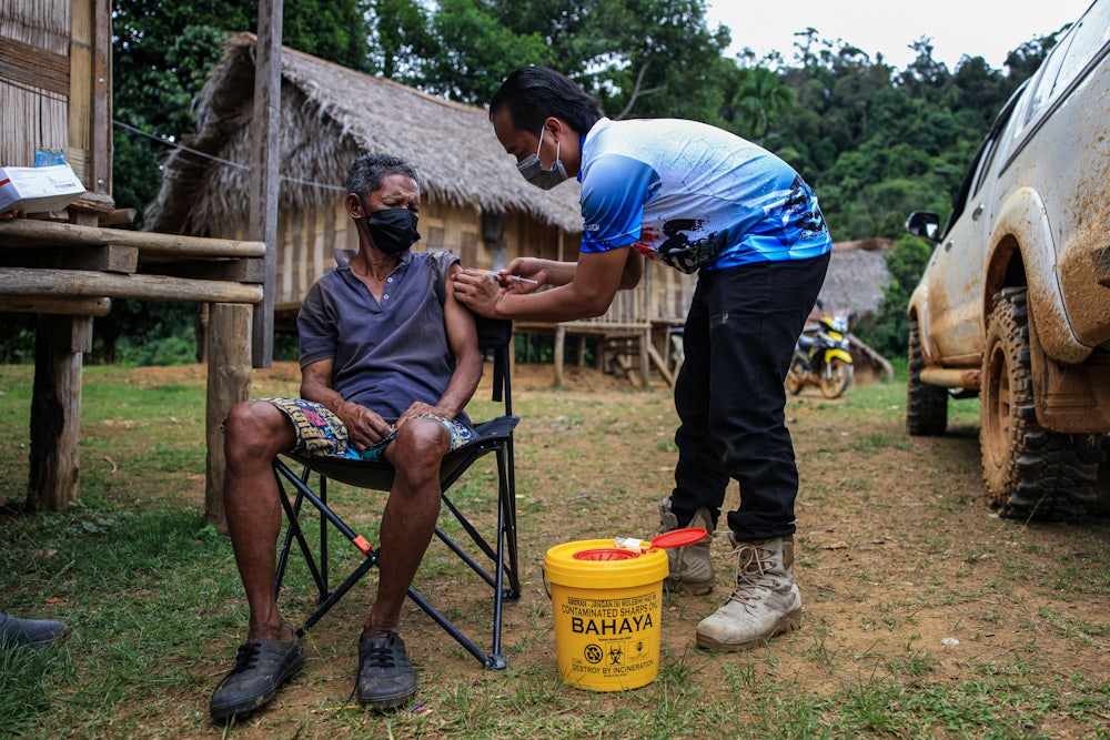 A health worker injects a Covid vaccine into a seated man's arm, outdoors, in front of a hut.