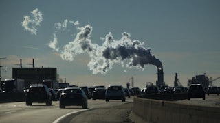 Cars move along a highway with a smokestack in the background.