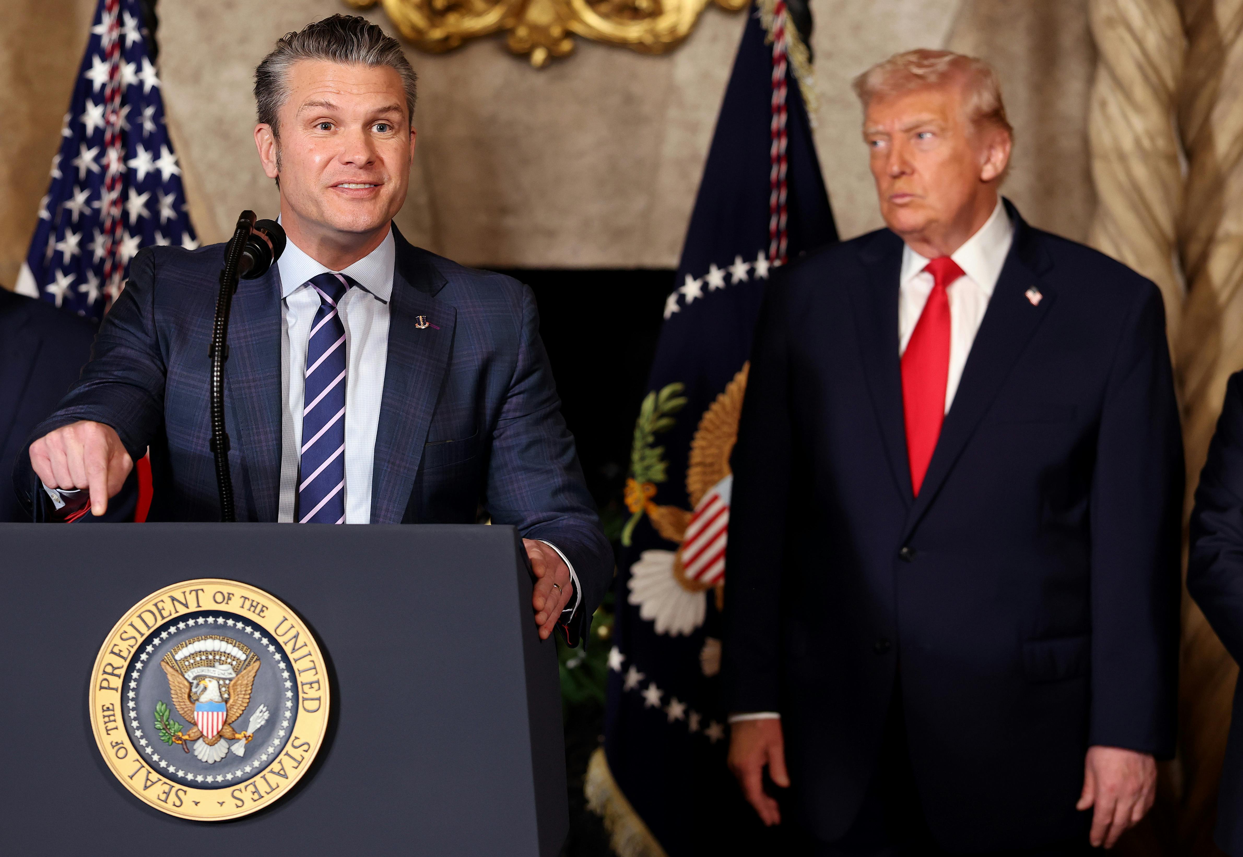 Defense Secretary Pete Hegseth gestures and speaks at a podium while Donald Trump stands behind him, watching