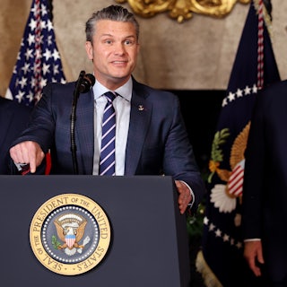 Defense Secretary Pete Hegseth gestures and speaks at a podium while Donald Trump stands behind him, watching