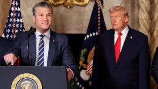 Defense Secretary Pete Hegseth gestures and speaks at a podium while Donald Trump stands behind him, watching