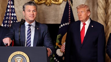 Defense Secretary Pete Hegseth gestures and speaks at a podium while Donald Trump stands behind him, watching