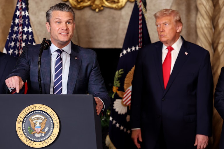 Defense Secretary Pete Hegseth gestures and speaks at a podium while Donald Trump stands behind him, watching