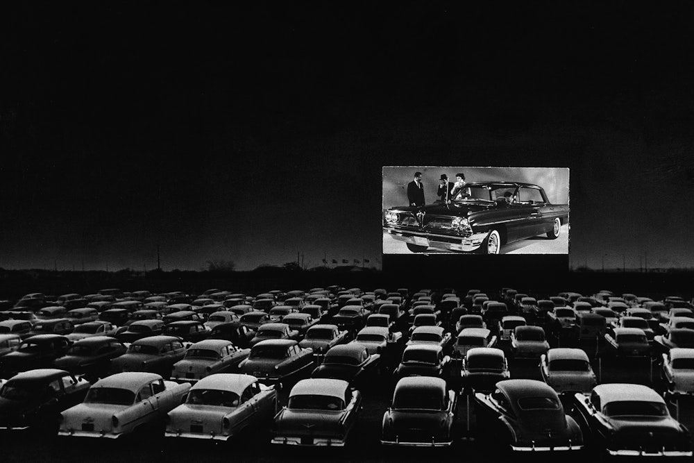 Vehicles fill a drive-in theater while people on the screen stand near a new car.