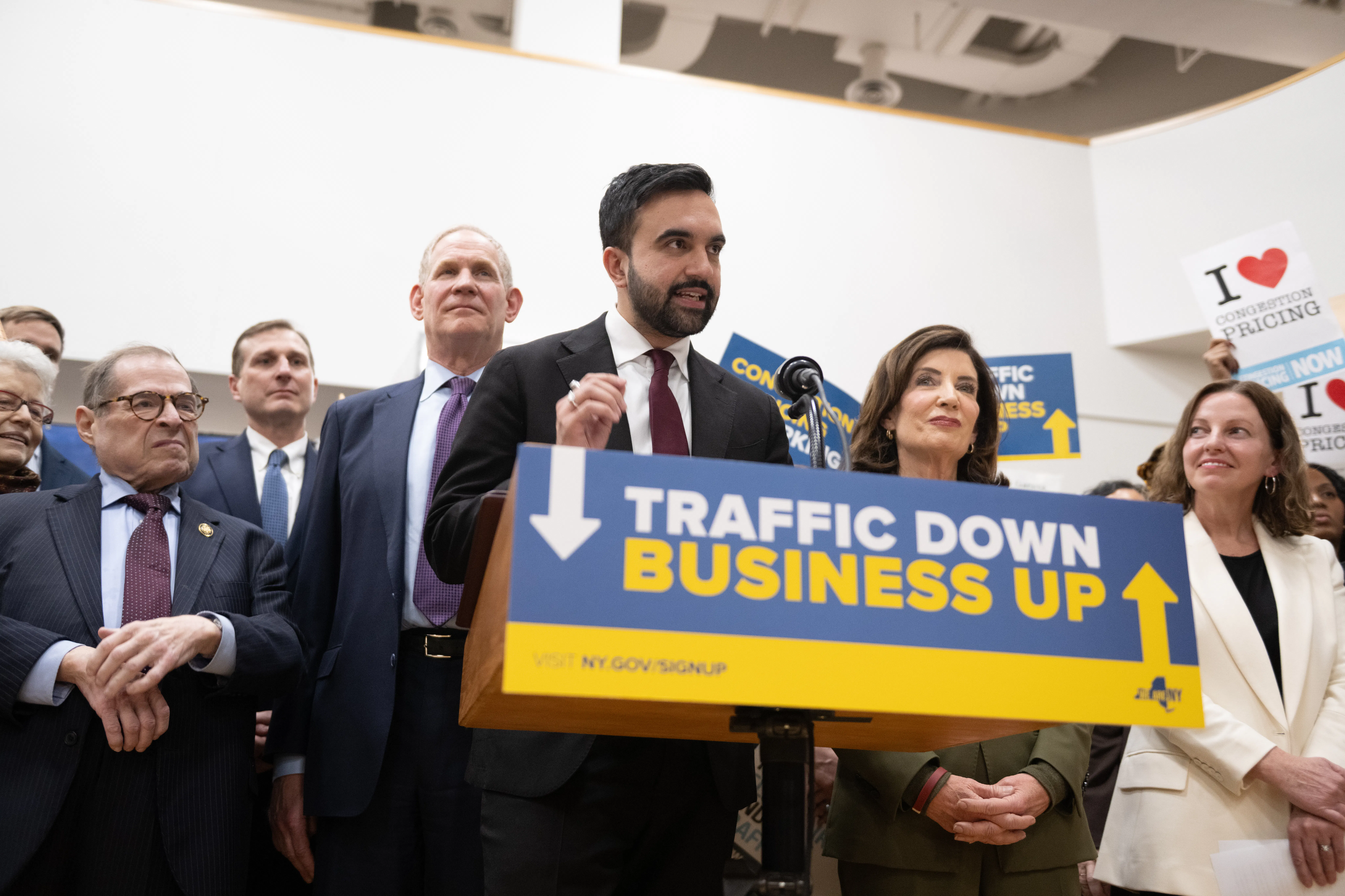 Zohran Mamdani, Kathy Hochul, and several others stand behind a podium reaching "Traffic Down. Business Up."
