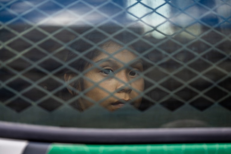 A small brown child sits in the back of a Border Patrol car. She is pictured through the Xed out rear window.