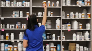 A woman takes a bottle off the top shelf of medications in a pharmacy