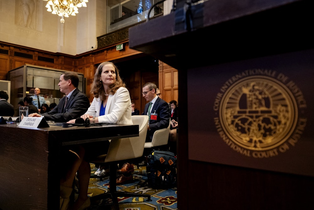 This photo, looking up from behind a lectern, shows a woman in a white blazer seated at a desk with a man in a suit to her right, with others in the room.