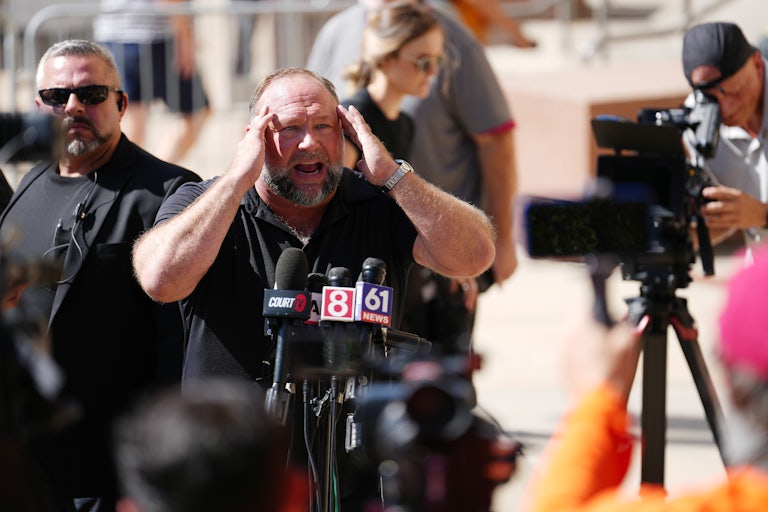 Alex Jones puts both hands on his head as if in distress. Press surrounds him.