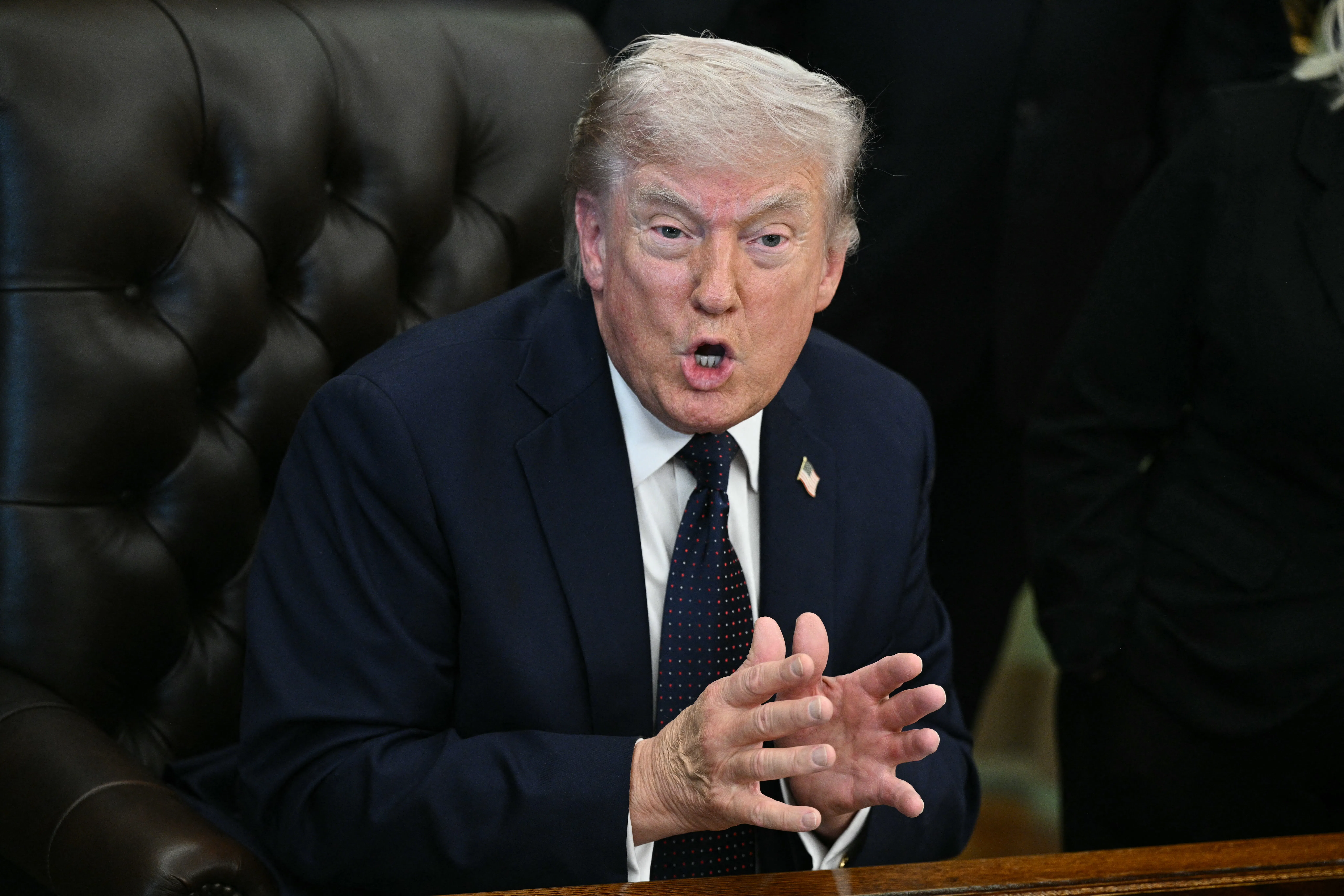 Donald Trump leans forward while sitting in his desk chair in the Oval Office. He holds both hands in front of him, palms almost touching, and speaks.