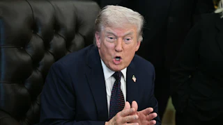 Donald Trump leans forward while sitting in his desk chair in the Oval Office. He holds both hands in front of him, palms almost touching, and speaks.