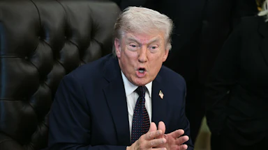 Donald Trump leans forward while sitting in his desk chair in the Oval Office. He holds both hands in front of him, palms almost touching, and speaks.