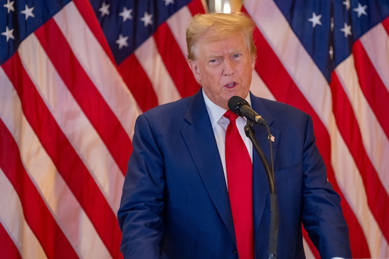 Donald Trump makes a weird face while speaking before a mic. Several U.S. flags are behind him.