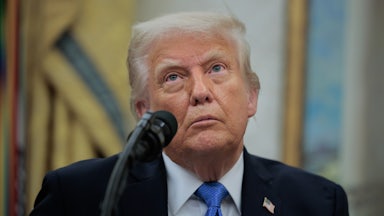 Donald Trump is staring into the upper left corner, wearing a dark suit and blue tie, with a microphone positioned to the left of his mouth.