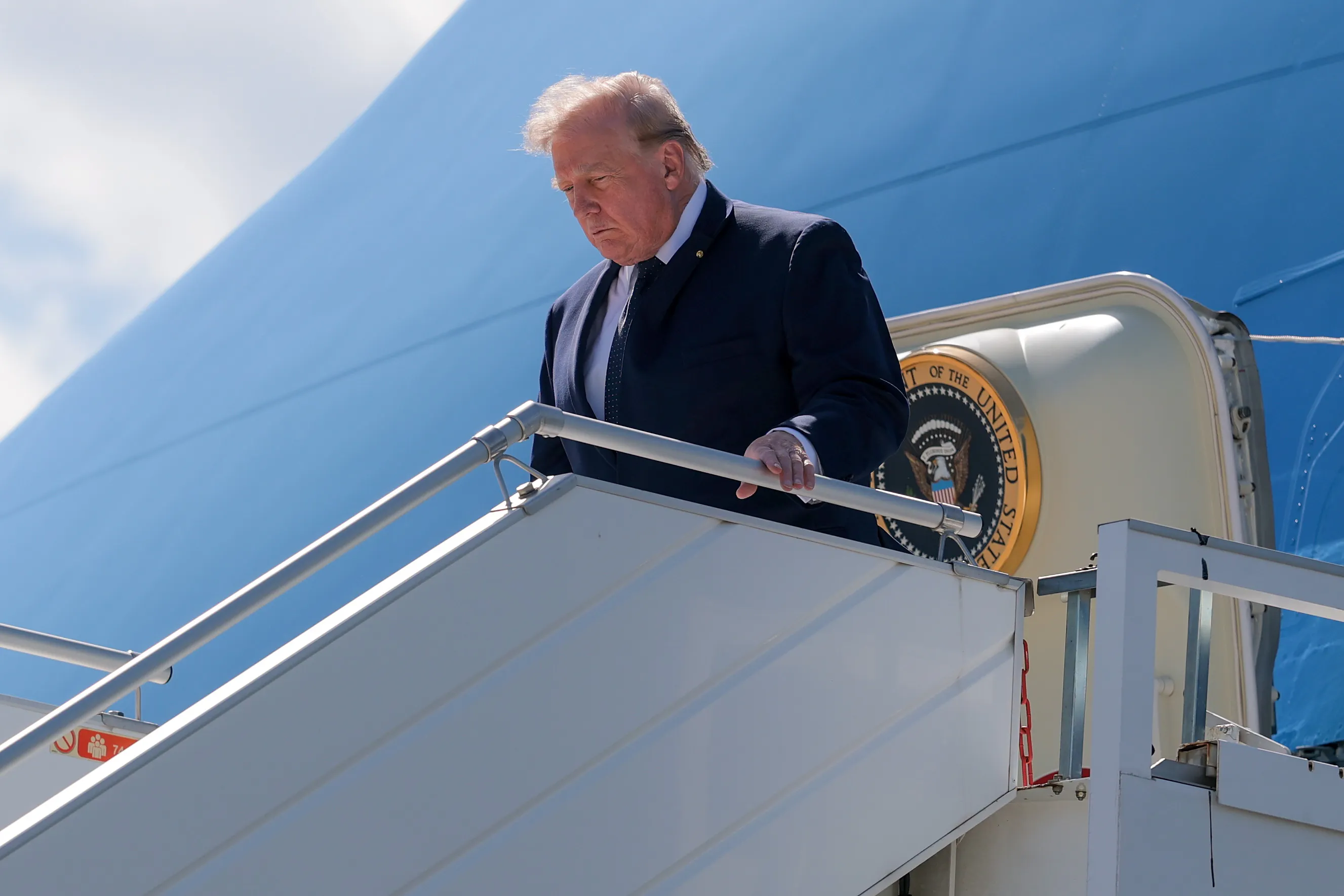 Donald Trump looks down and holds the railing while disembarking from Air Force One