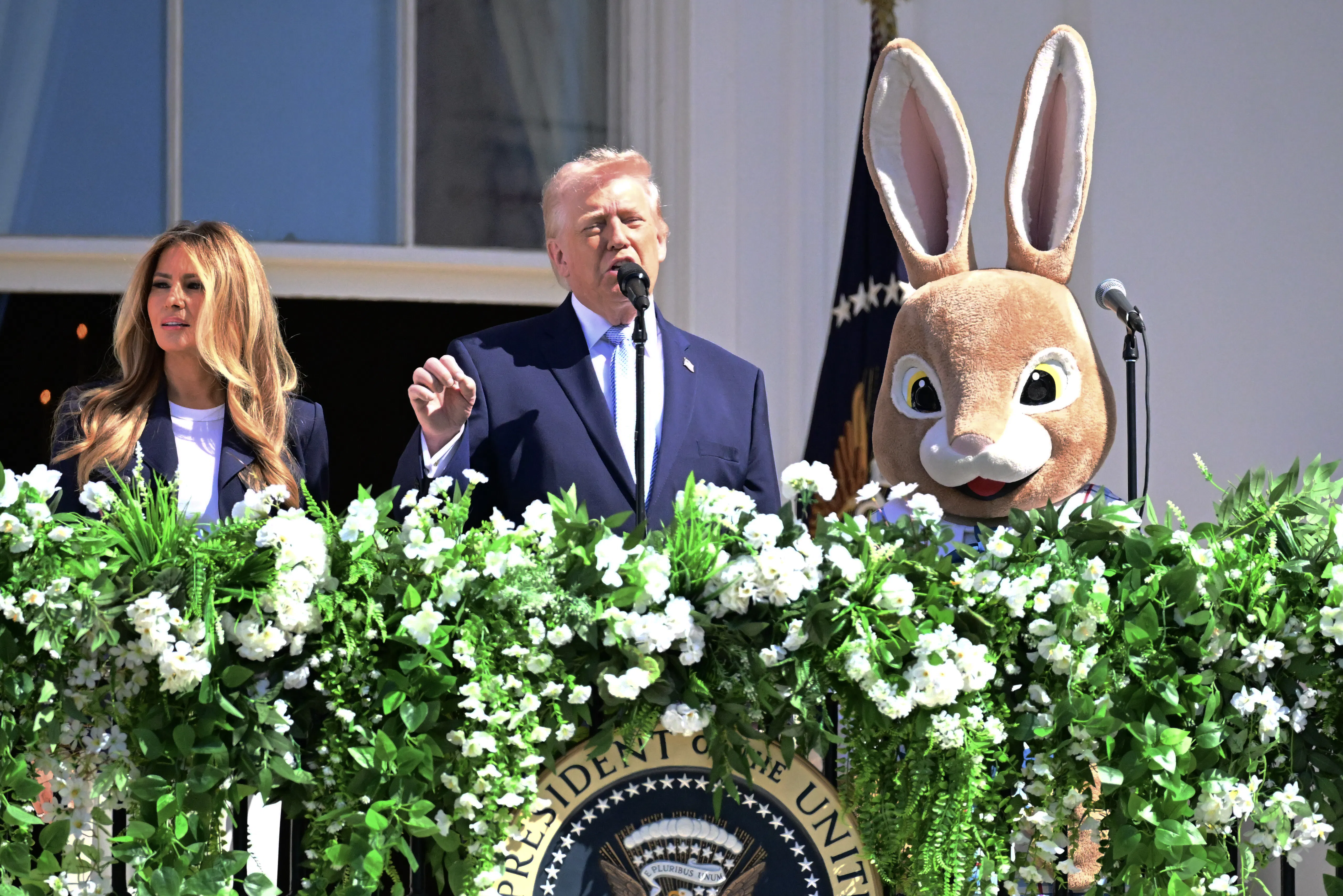 Donald Trump gestures and speaks while standing on the White House balcony. He is flanked by Melania Trump and a person in an Easter Bunny costume