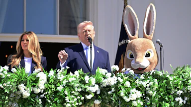 Donald Trump gestures and speaks while standing on the White House balcony. He is flanked by Melania Trump and a person in an Easter Bunny costume