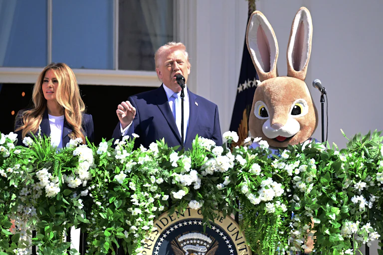 Donald Trump gestures and speaks while standing on the White House balcony. He is flanked by Melania Trump and a person in an Easter Bunny costume