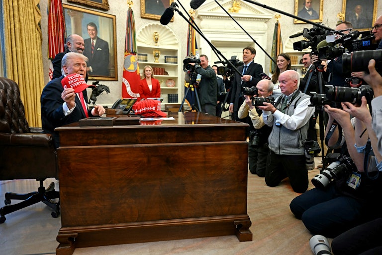 Donald Trump is seated at his desk in the Oval Office holding up his new MAGA cap for more than a dozen reporters holding cameras and mics. There is a pile of red caps sitting on his desk. Howard Lutnick and Karoline Leavitt look on in the background and smile.