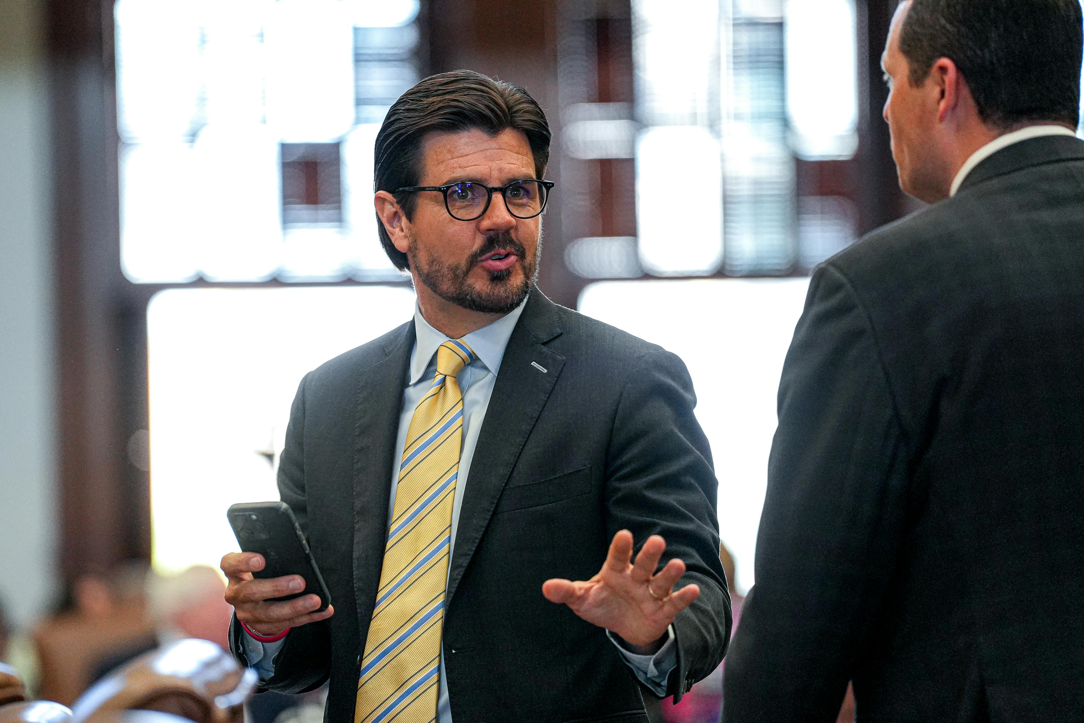 Texas state Representative Cole Hefner speaks while standing in the state Capitol. He holds a cell phone in one hand and gestures with the other
