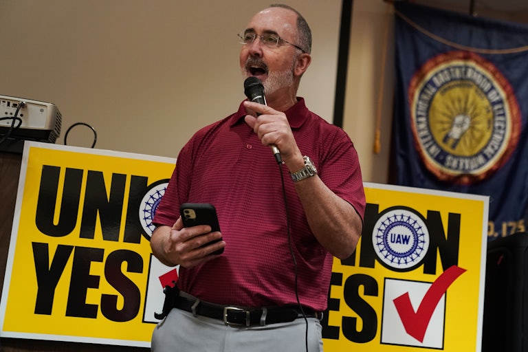 Shawn Fain speaks with a mic in his hand. He stands in front of a sign that refers to unions and has a "Yes" checkbox marked.
