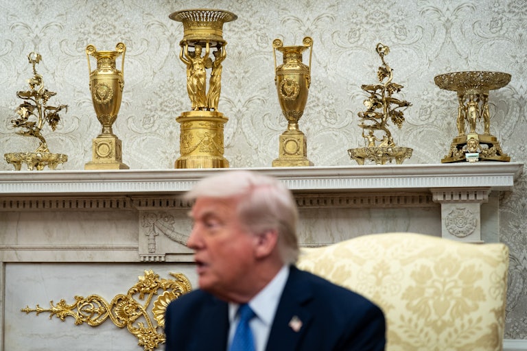 President Donald Trump sits in front of a row of golden vases and trophies.