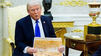 Donald Trump sits in the Oval Office and speaks while holding up a rendering of the gilded ballroom he's building at the White House