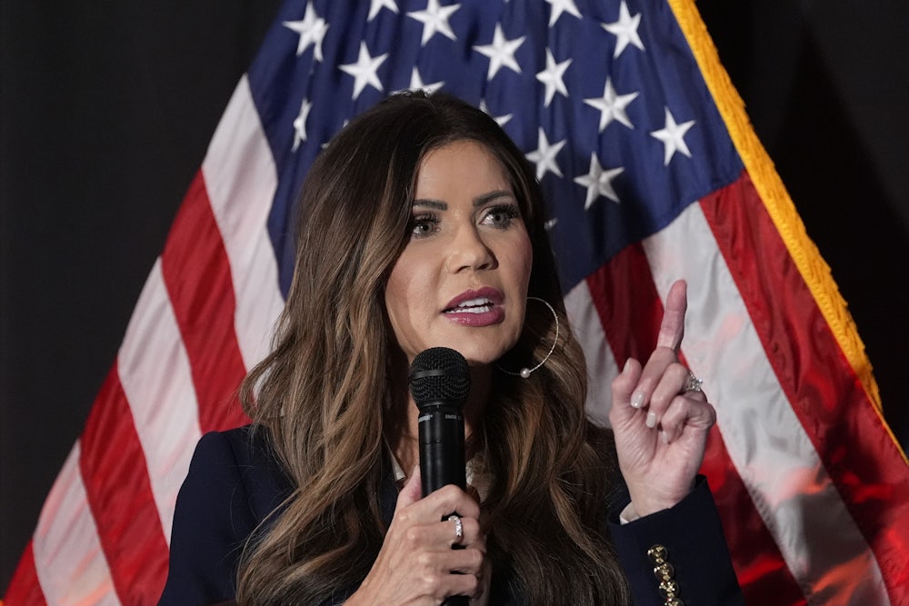 Kristi Noem holds up a finger while speaking into a microphone as she stands in front of an American flag, against a dark background.
