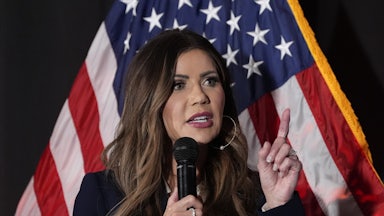 Kristi Noem holds up a finger while speaking into a microphone as she stands in front of an American flag, against a dark background.