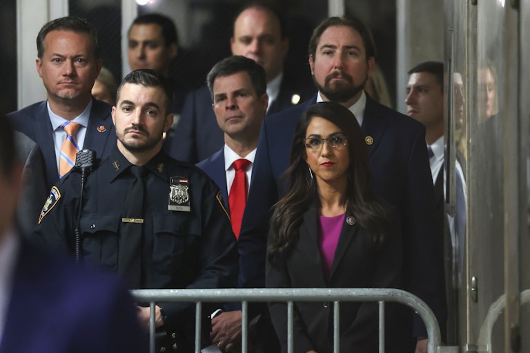 Lauren Boebert stands in a crowd behind a metal barricade. A security guard stands to the left of her.