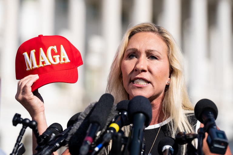 Marjorie Taylor Greene speaks in the sun with several mics before her. She holds a red Make America Great again cap in her hand and looks angry.