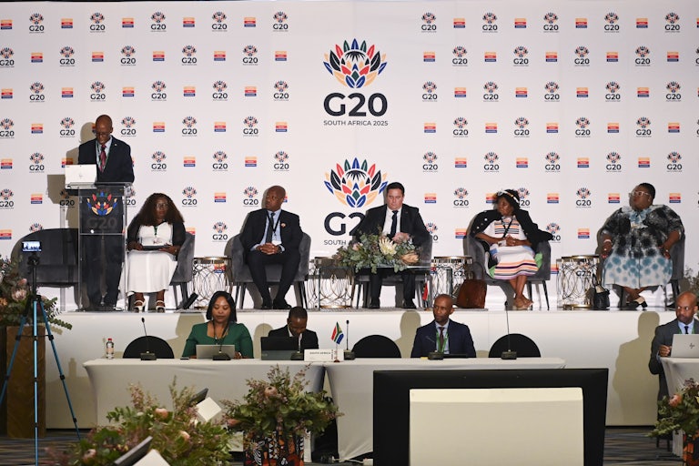 Several Black people sit on a dias in front of a large backdrop that says G20 South Africa 2025. They listen to a man speaking behind a lectern next to them.