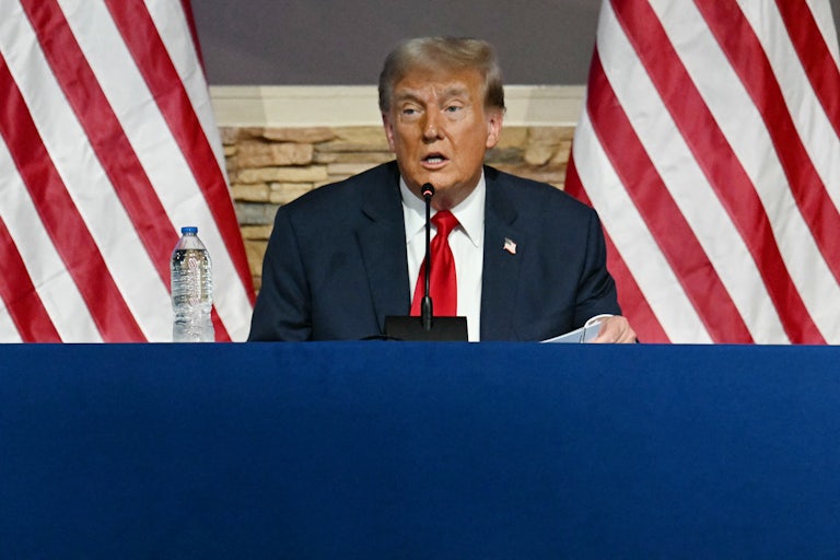 Donald Trump is seated at a blue table and he looks confused. He is seated in the middle of two large U.S. flags behind him.