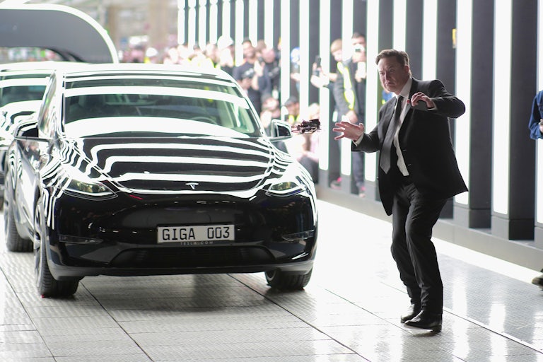 Elon Musk strikes an odd pose while standing next to Tesla vehicles while wearing a suit.
