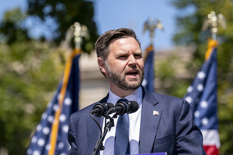 J.D. Vance speaking at a lectern outdoors. A row of U.S. flags are behind him.