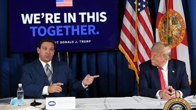 Ron DeSantis and Donald Trump sit at a table with a banner reading "We're in this together" behind them.